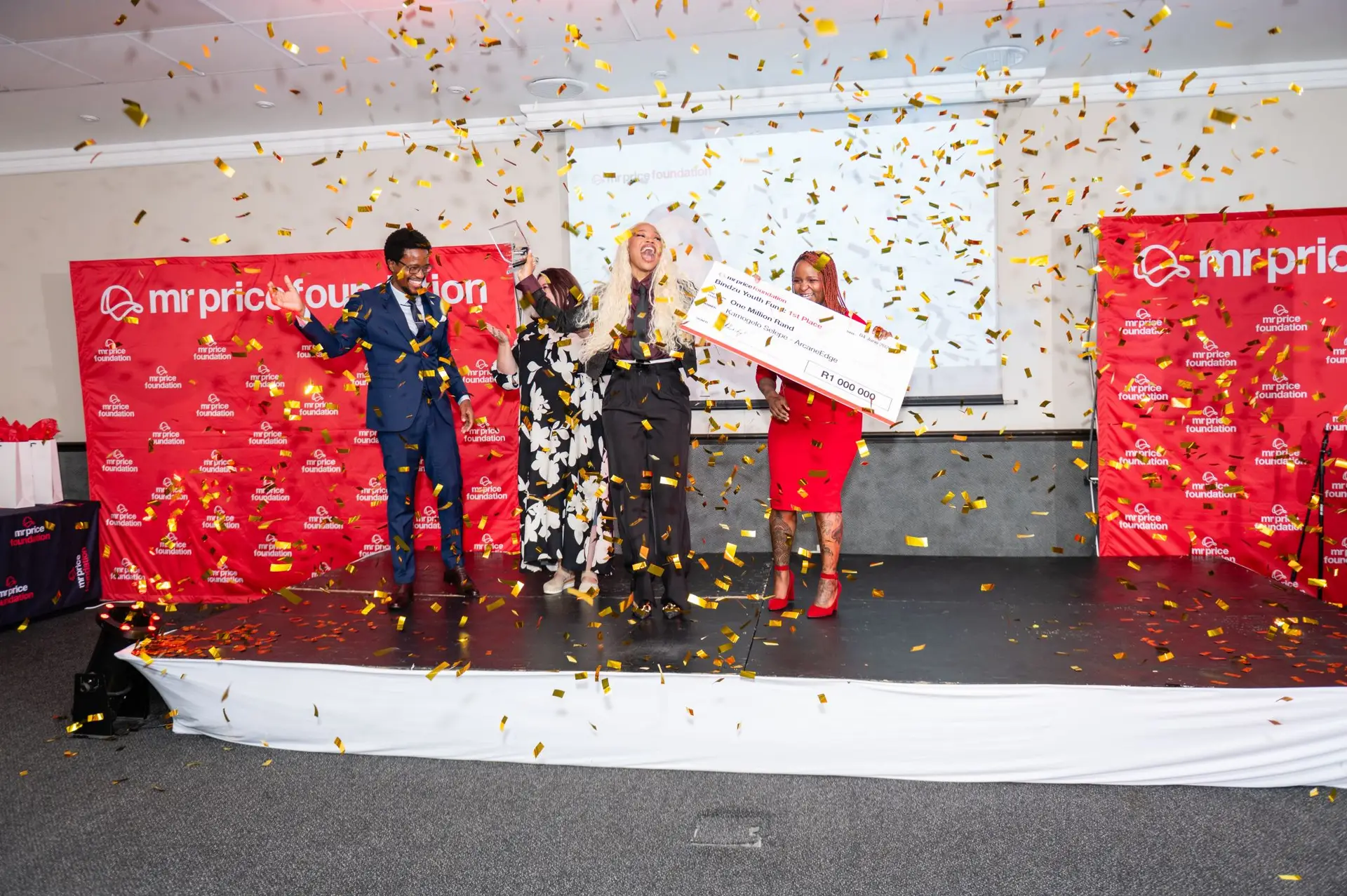 Group of four people celebrating on stage as gold confetti falls; one person holds a large ceremonial check for 1,000,000 rand with a Mr Price Foundation backdrop.
