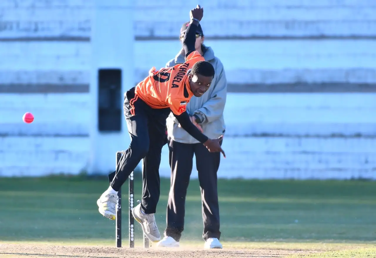 Cricketer in an orange jersey delivering a ball near the stumps, with a pink ball in midair and an official in the background far behind.