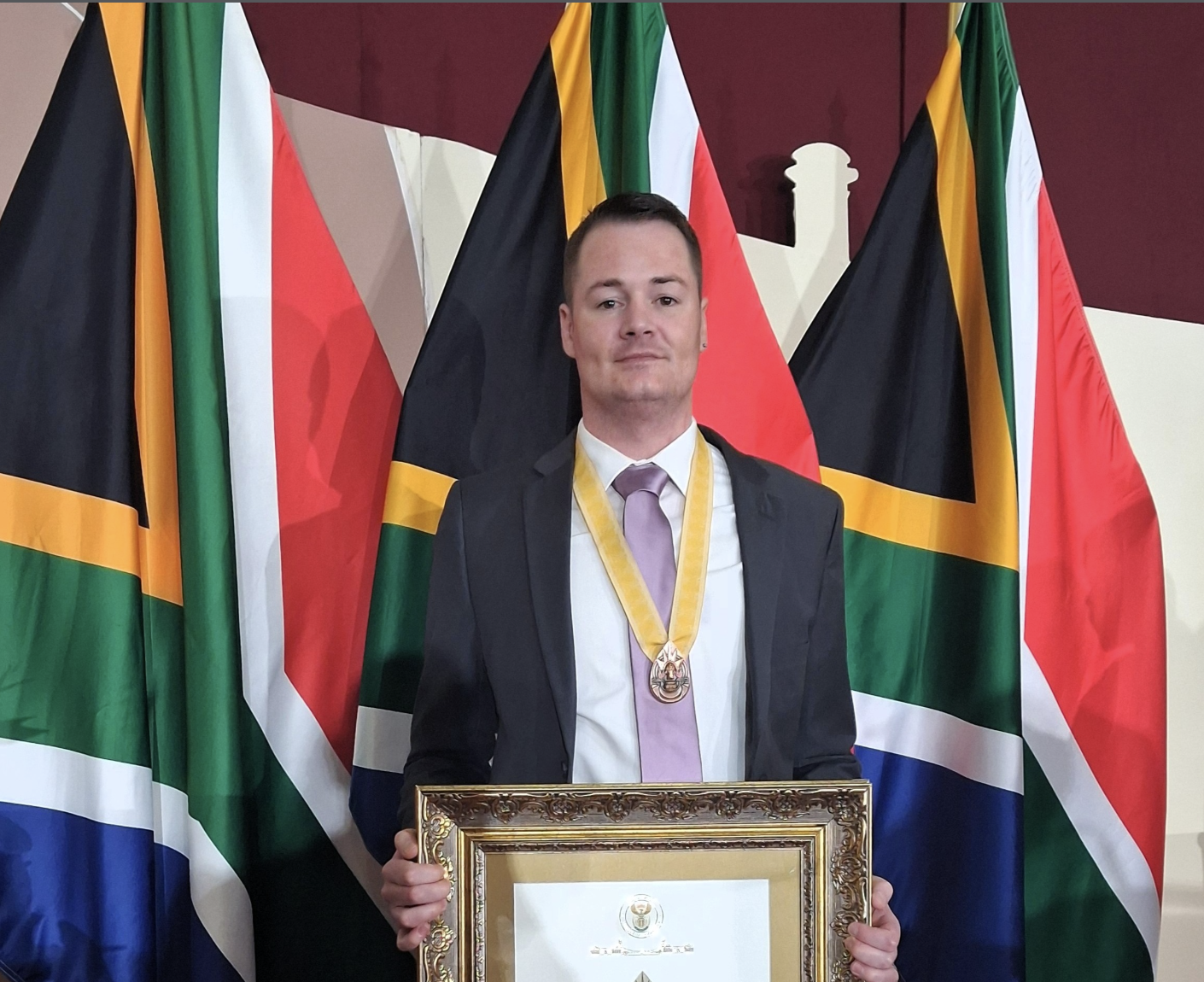 Man in a suit with a gold medal around his neck, holding a framed certificate at an award ceremony with multiple flags in the background.