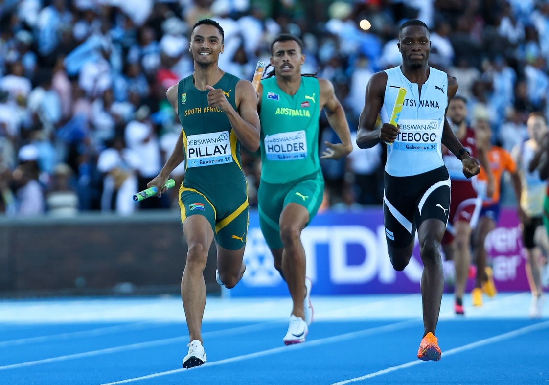 Three male sprinters run side by side on a bright blue track during a race, colorful bibs visible on their chests and a cheering crowd in the background.