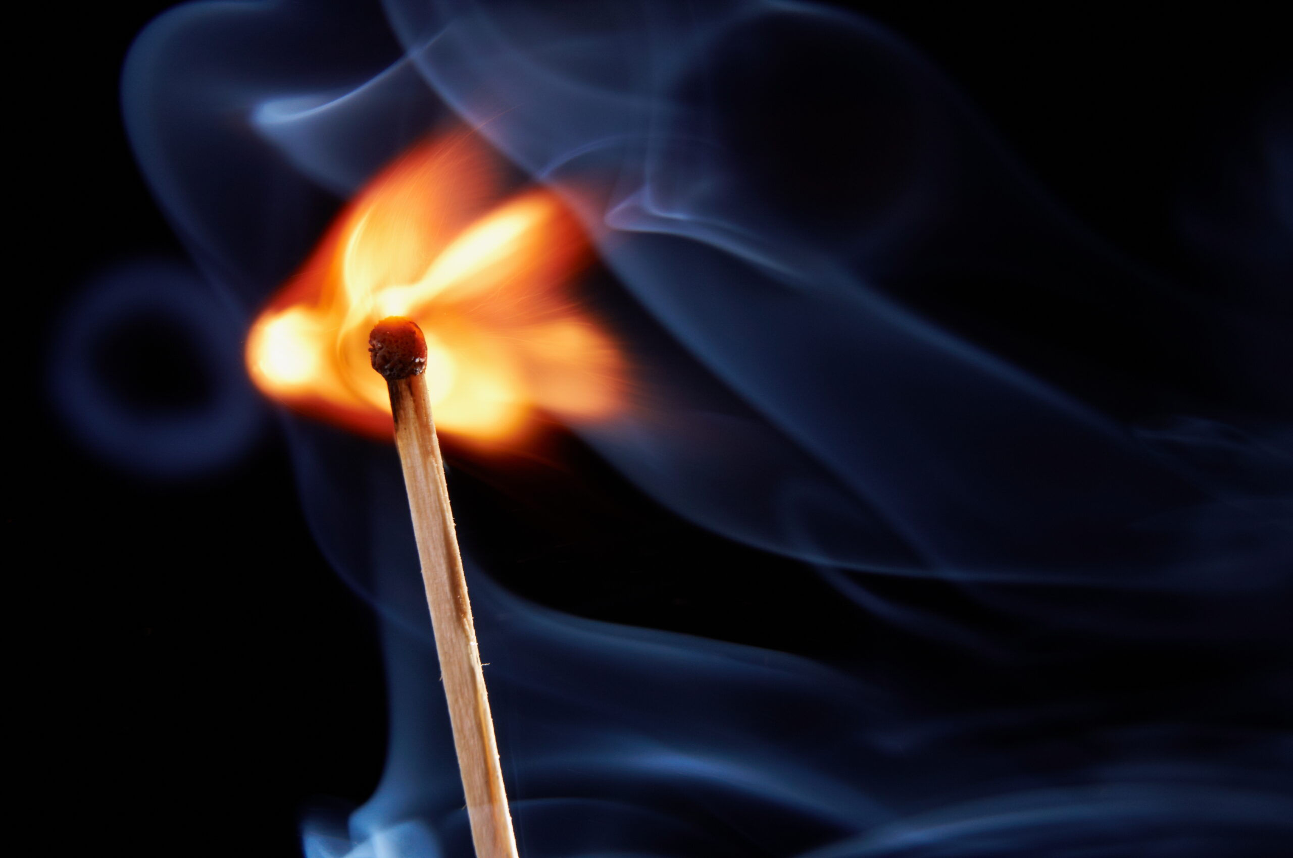Close-up of a lit match with a bright flame and curling blue smoke against a dark background, focusing on the match head.