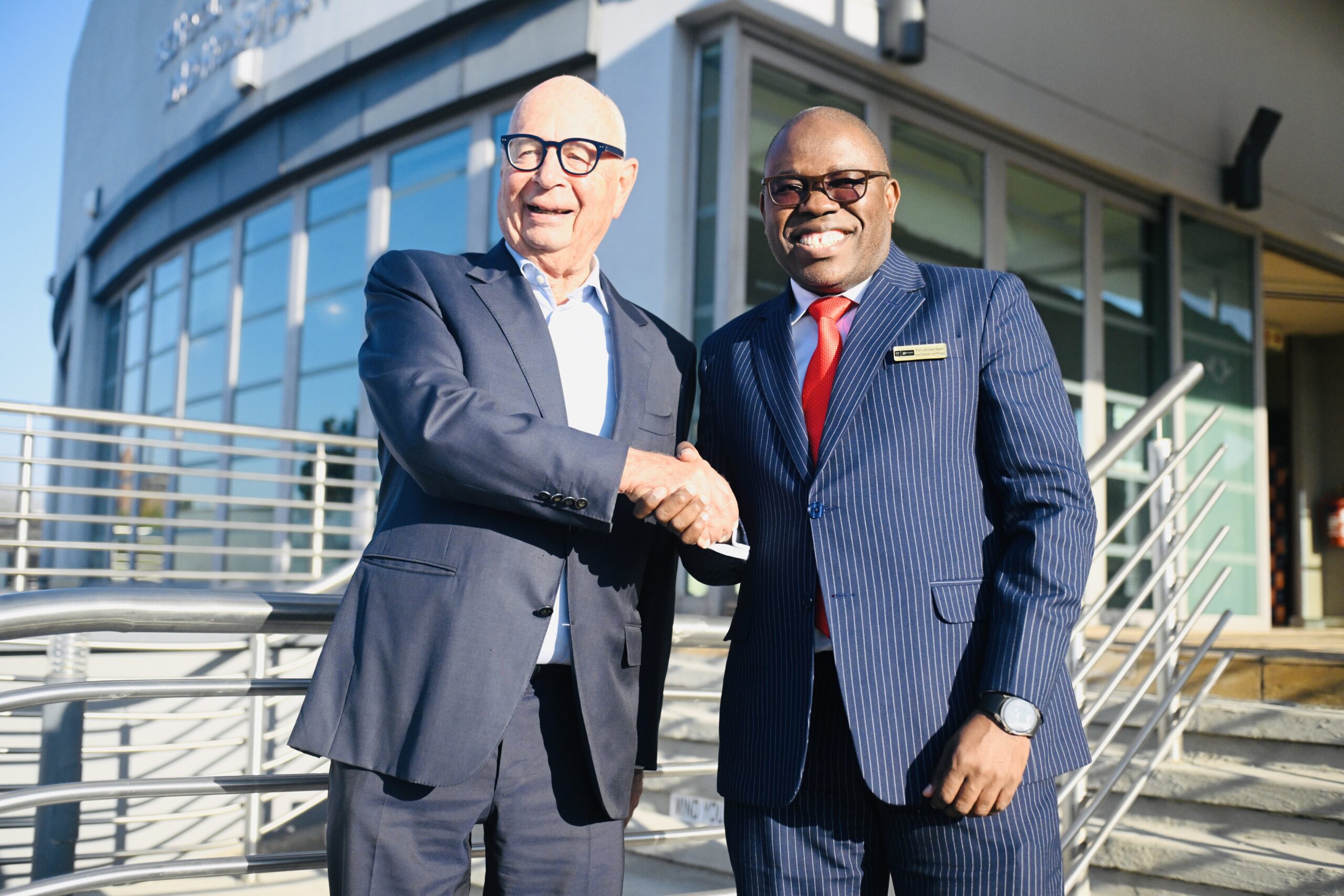 Two businessmen in suits shake hands outside a modern building with glass windows and metal railings, smiling at the camera.