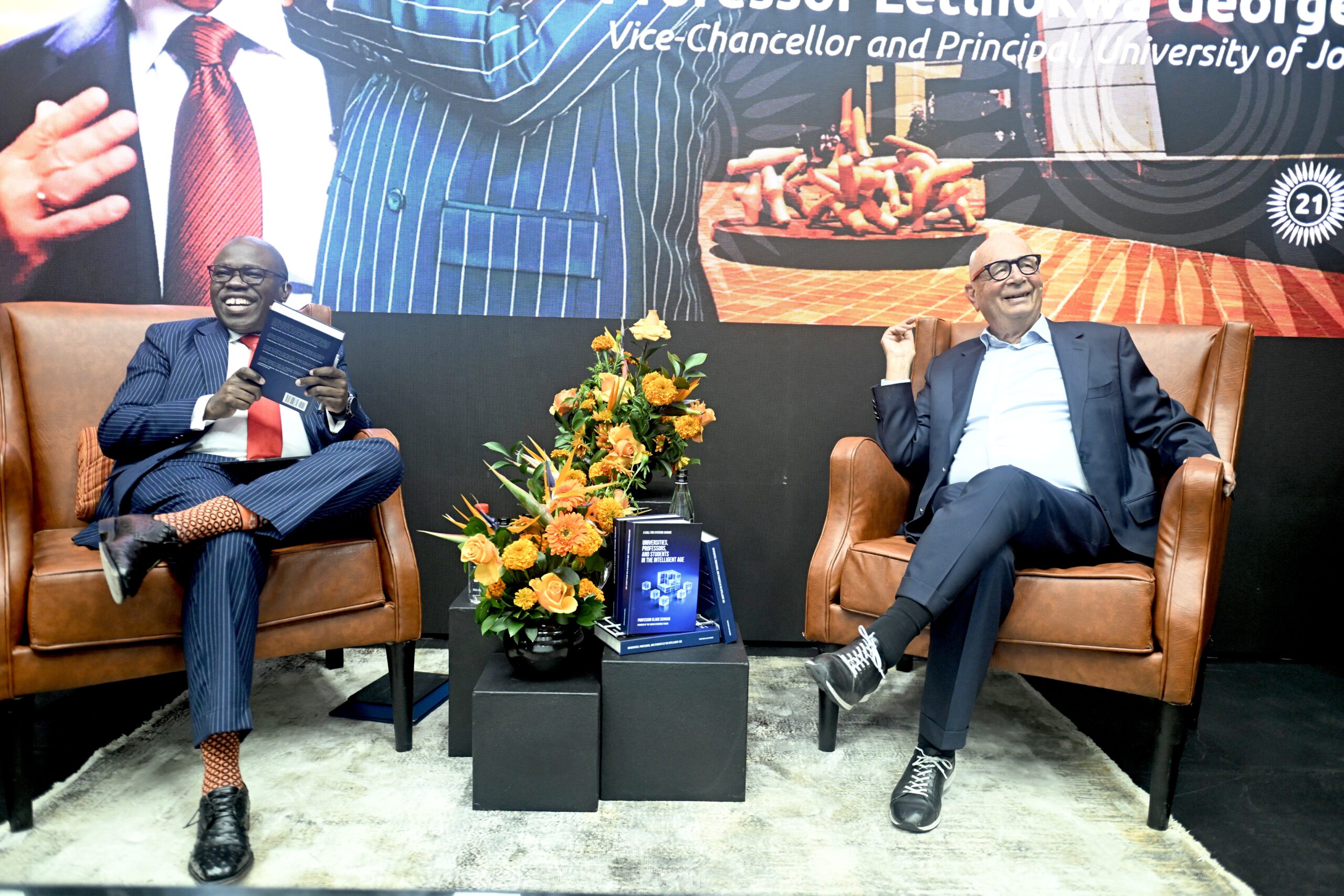 Two men in suits seated on brown leather chairs during a panel discussion, smiling, with a flower arrangement and books on a black table in front.