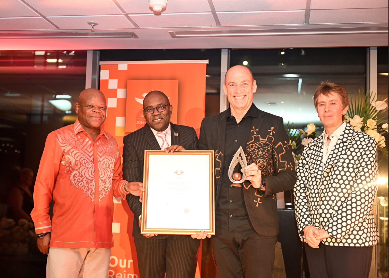 Four people at an awards ceremony pose with a framed certificate and a glass trophy against an orange backdrop.